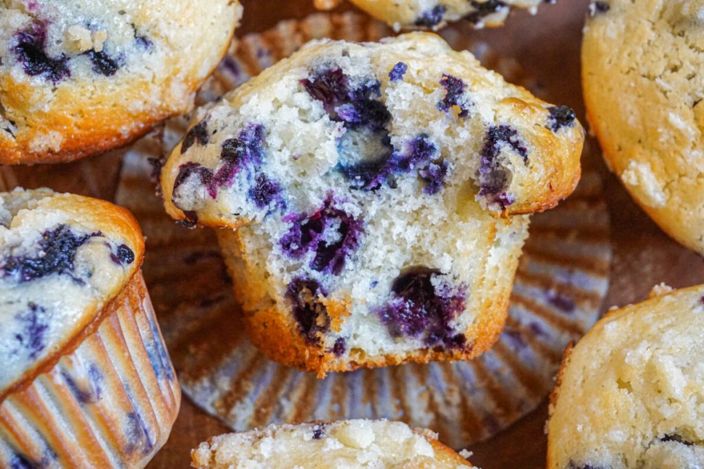 A close-up of blueberry muffins, with one muffin halved to show its moist interior filled with blueberries, resting on a brown muffin liner.