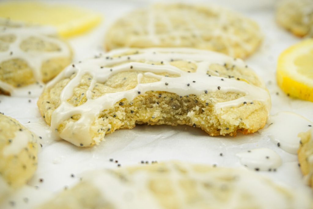 A lemon poppy seed cookie with white icing and a bite taken out, surrounded by more cookies and lemon slices on a white surface.