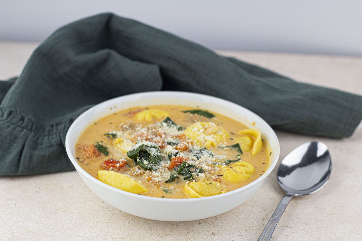 A bowl of creamy soup with tortellini, spinach, and tomatoes, topped with grated cheese. A spoon lies next to the bowl and a dark green cloth is in the background.
