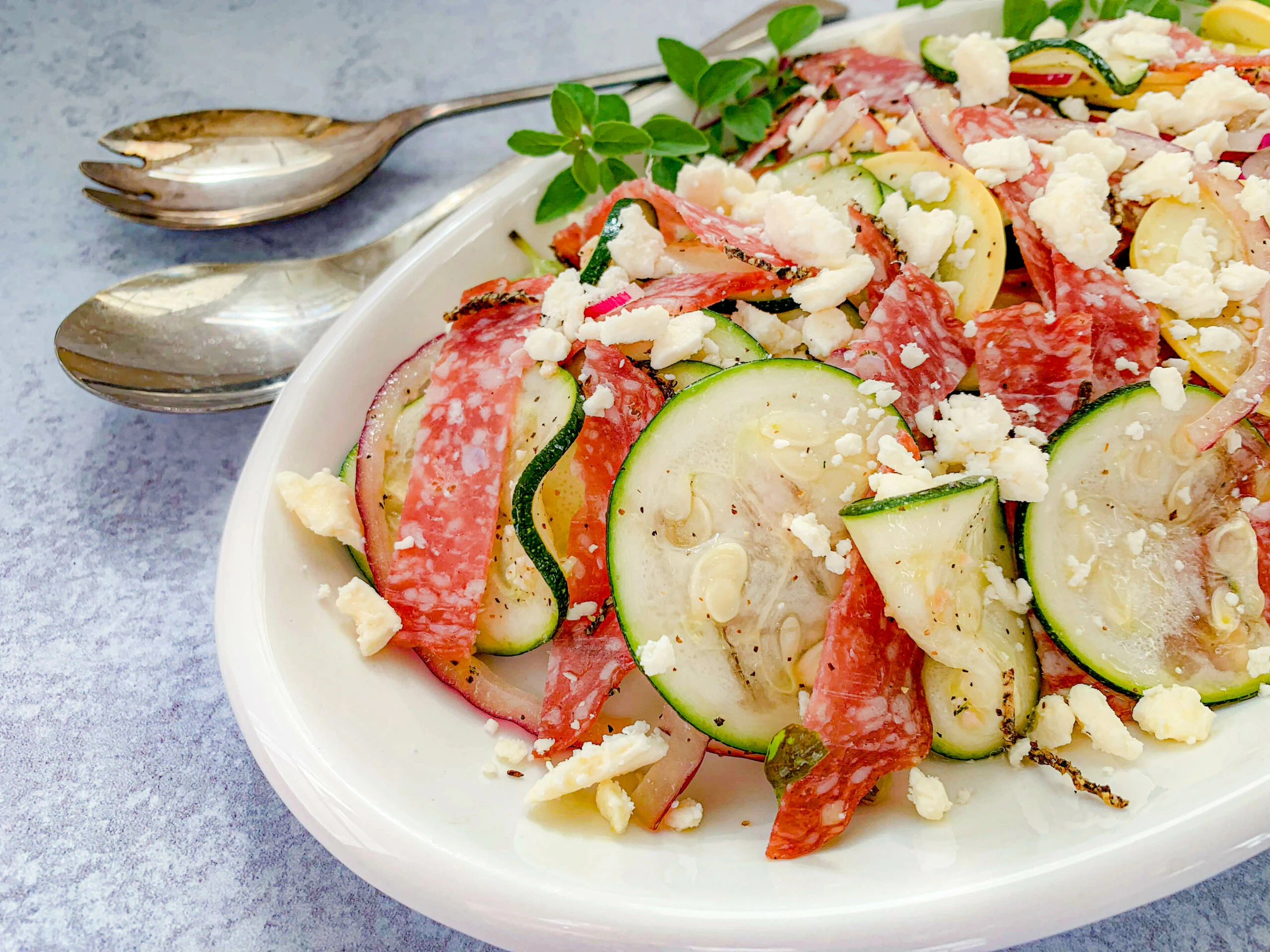 A white plate with a salad of sliced zucchini, yellow squash, salami, crumbled cheese, and herbs, with serving utensils beside it.