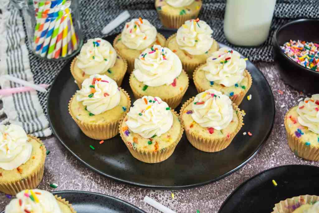 A plate of funfetti cupcakes with white frosting and colorful sprinkles, surrounded by more cupcakes, a bottle of milk, and a bowl of sprinkles.