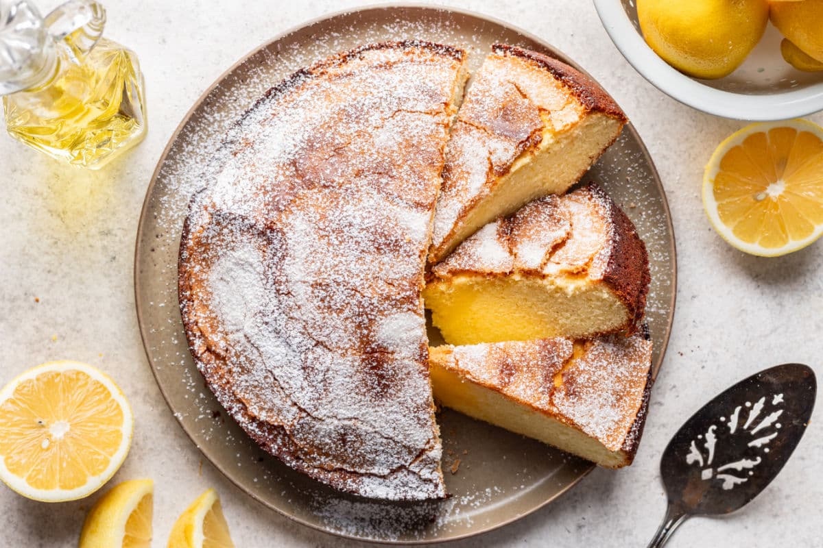 A round lemon cake topped with powdered sugar sits on a plate, with three slices cut. Whole and sliced lemons and a cake server are nearby.