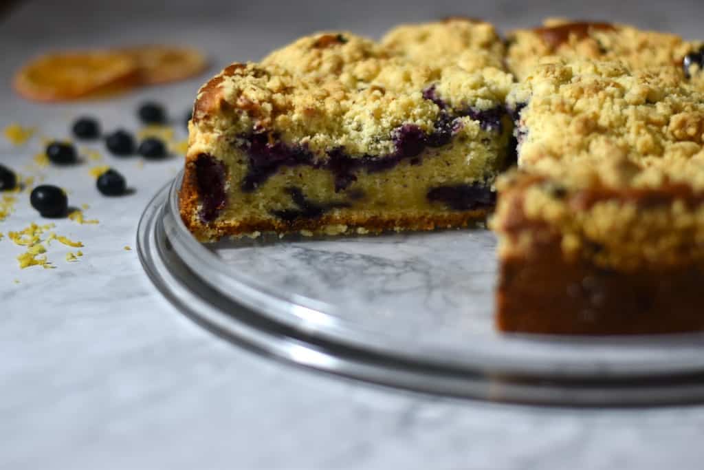 A round blueberry crumb cake on a glass plate with a slice removed; blueberries and orange zest are scattered in the background.