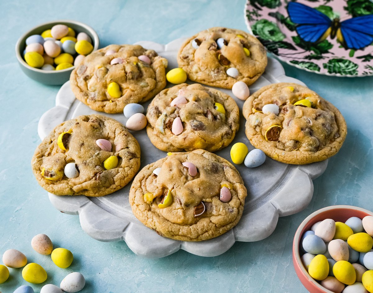 Six cookies with colorful candy-coated chocolate pieces are arranged on a marble platter, surrounded by small bowls of similar candies on a light blue surface.