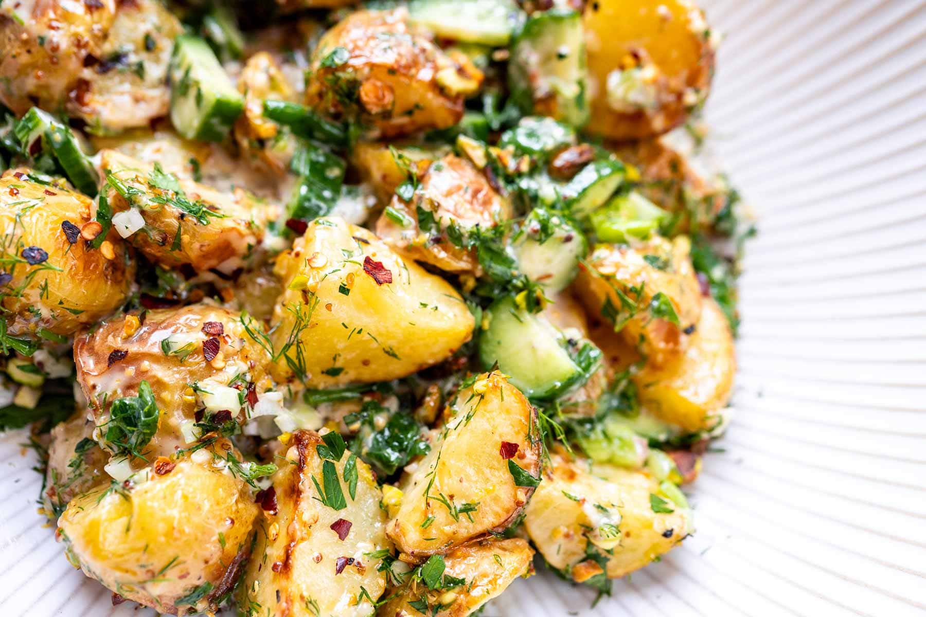 Close-up of roasted potato salad mixed with chopped herbs, cucumber, and a creamy dressing, served on a white plate.