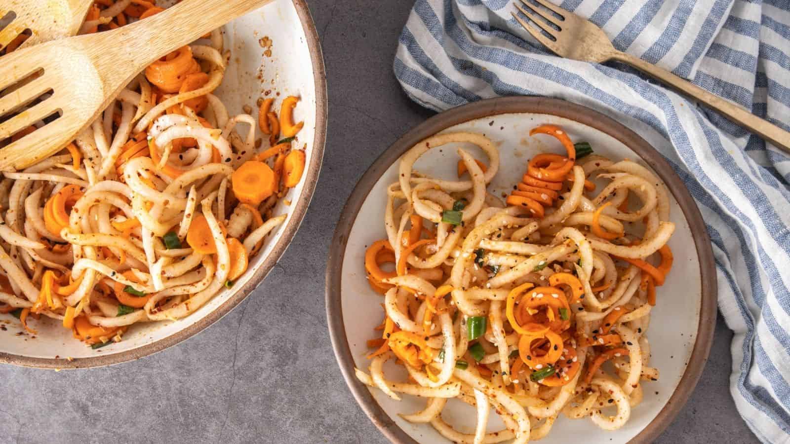 A plate and pan of stir-fried udon noodles with sliced carrots and green onions, served with wooden utensils on a gray surface next to a striped kitchen towel.