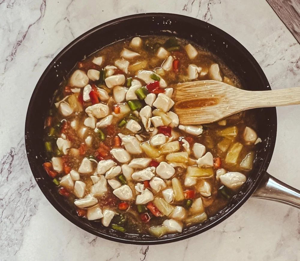 A skillet with diced chicken, chopped vegetables, and sauce being stirred with a wooden spatula on a light countertop.