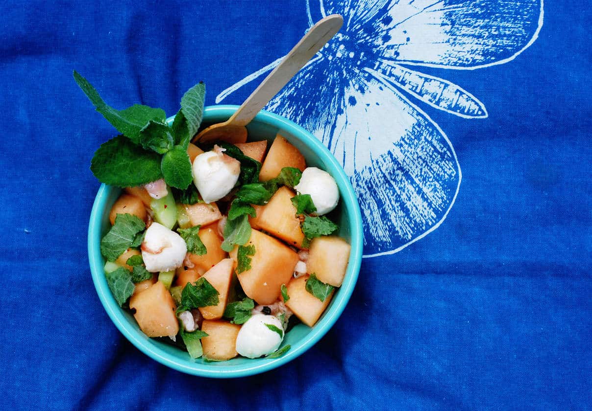 A bowl of melon salad with mozzarella balls and fresh mint leaves sits on a blue cloth with a white butterfly print.