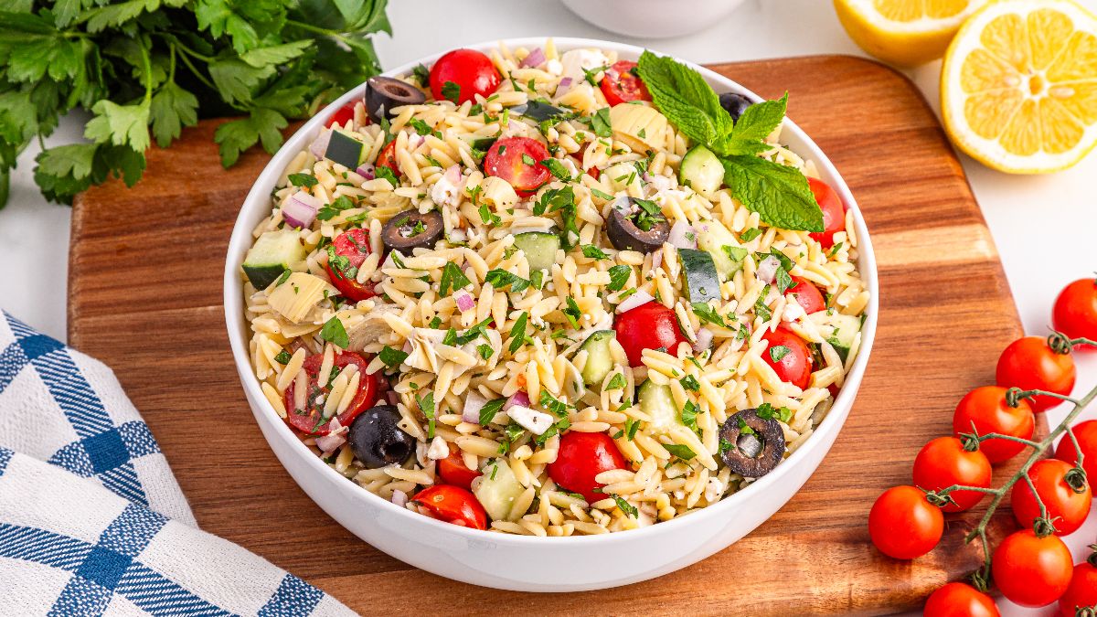 A bowl of orzo pasta salad with cherry tomatoes, cucumbers, black olives, red onion, artichoke hearts, parsley, and mint on a wooden board, with lemons and fresh vegetables nearby.