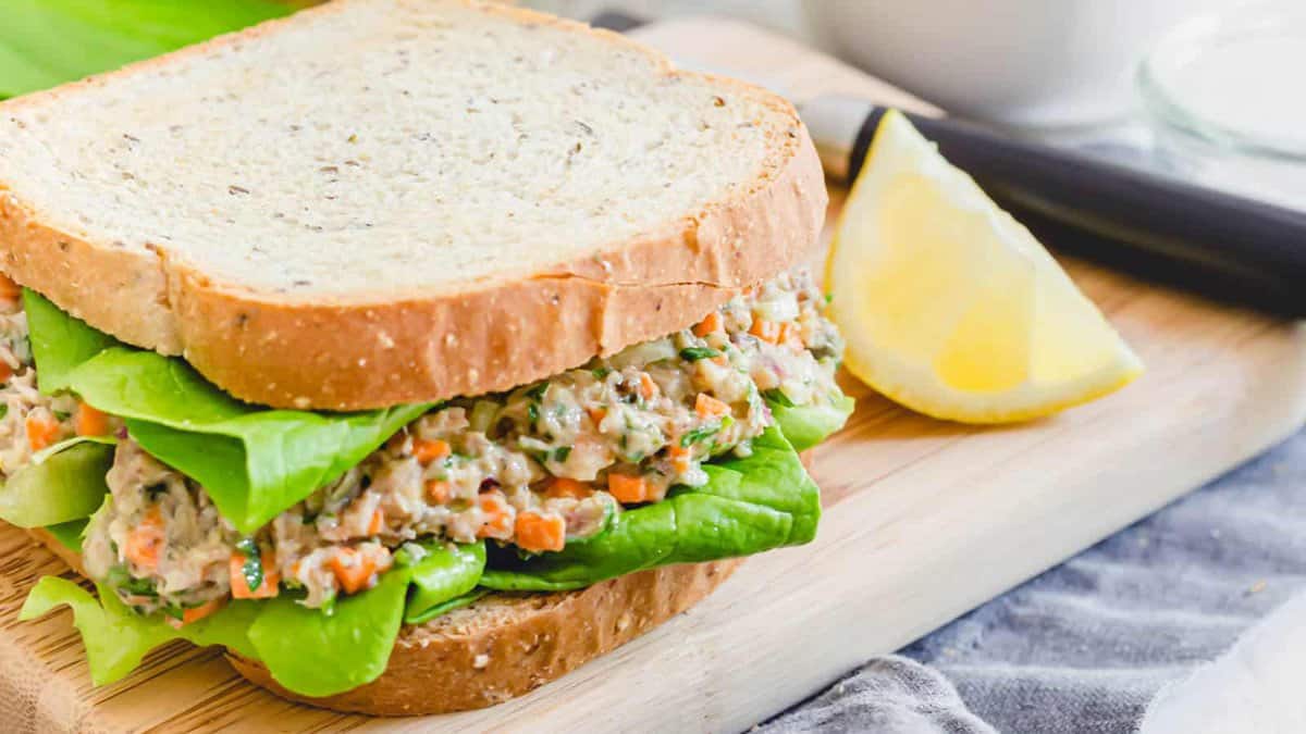 A tuna salad sandwich with lettuce on whole wheat bread sits on a wooden cutting board next to a lemon wedge and a knife.