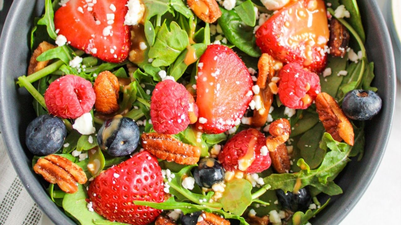 A salad bowl containing fresh spinach leaves, strawberries, raspberries, blueberries, pecans, crumbled cheese, and a light drizzle of dressing.
