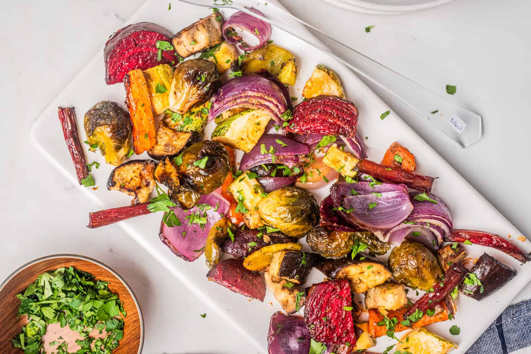 A white platter with roasted vegetables including carrots, red onions, Brussels sprouts, beets, and zucchini, topped with chopped fresh herbs.
