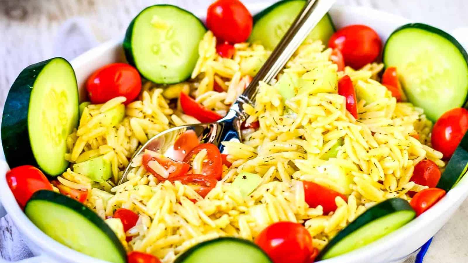 A bowl of orzo pasta salad with halved cherry tomatoes, cucumber slices, and a metal fork.