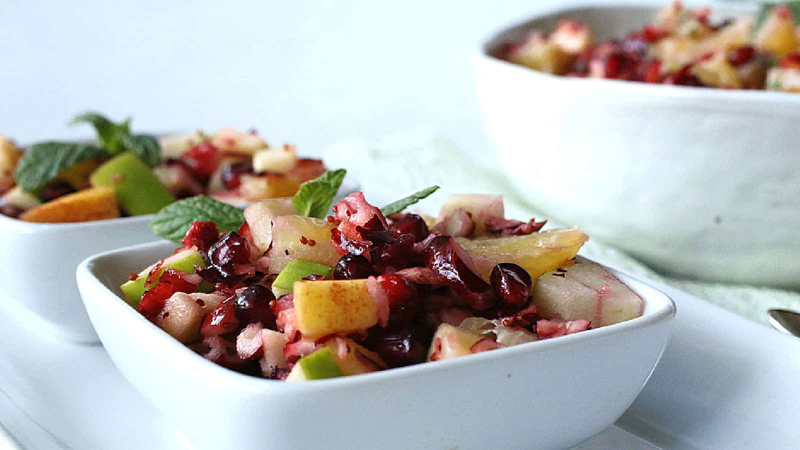 A close-up of a fruit salad with diced apples, pomegranate seeds, and mint leaves, served in small white bowls on a white surface.