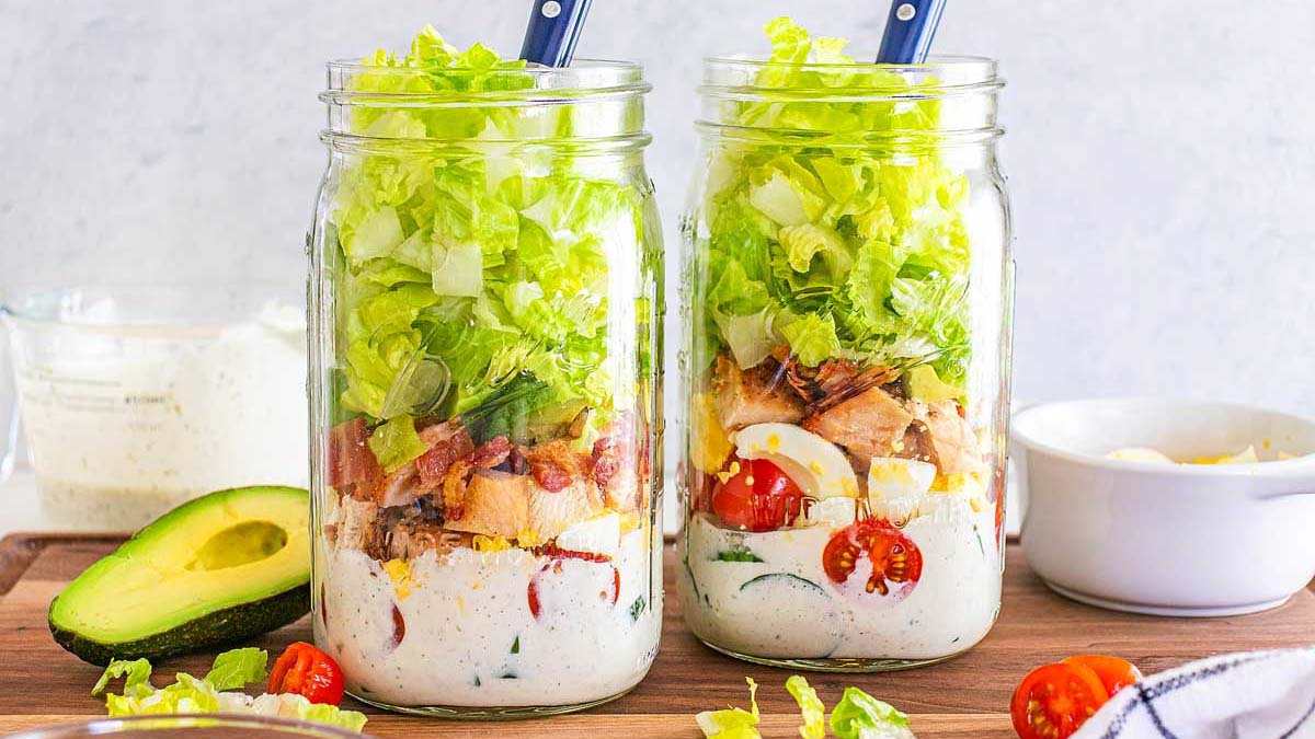 Two mason jars filled with layered salad ingredients including ranch dressing, cherry tomatoes, eggs, chicken, bacon, and chopped lettuce, each with a fork inside.