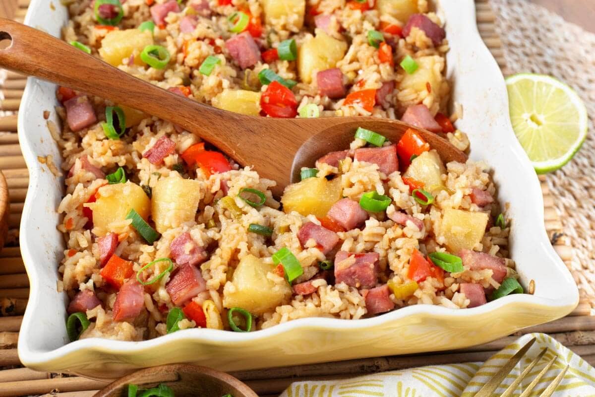 A casserole dish filled with fried rice, diced ham, pineapple chunks, red bell pepper, and green onions, with a wooden serving spoon and a half lime nearby.