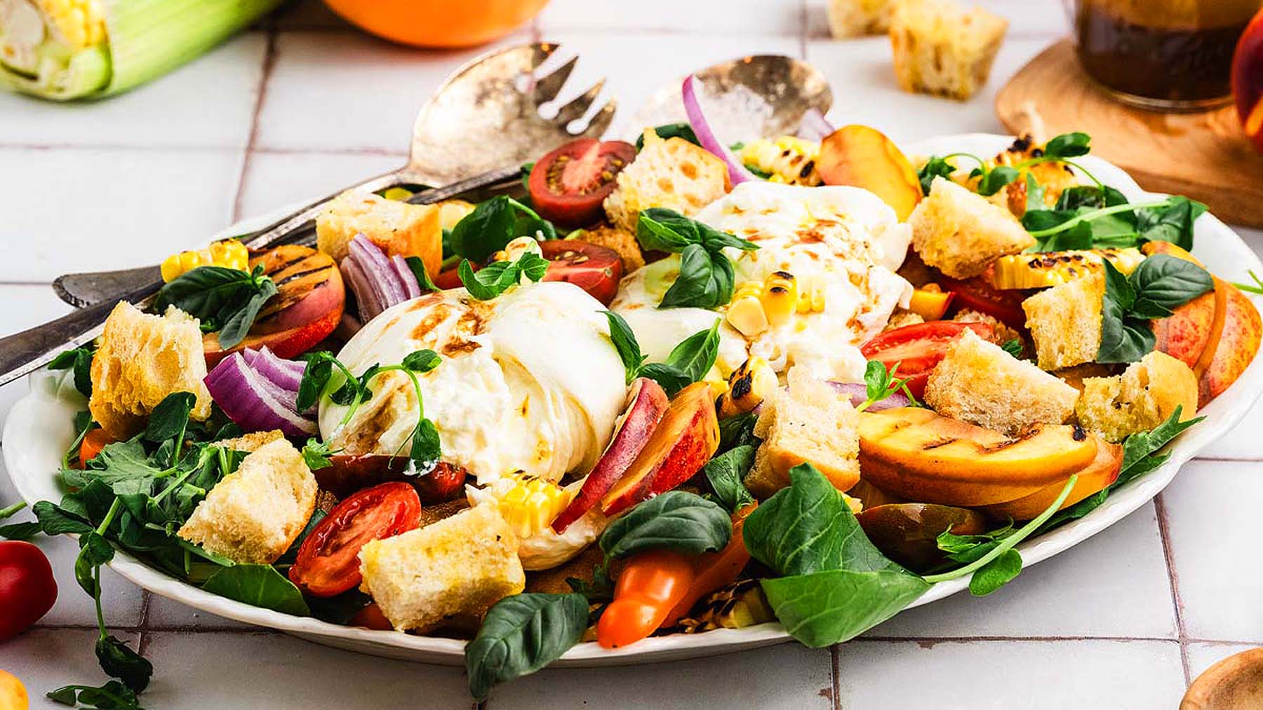 A plate of salad with grilled peaches, cherry tomatoes, red onions, leafy greens, croutons, and mozzarella cheese, garnished with herbs.