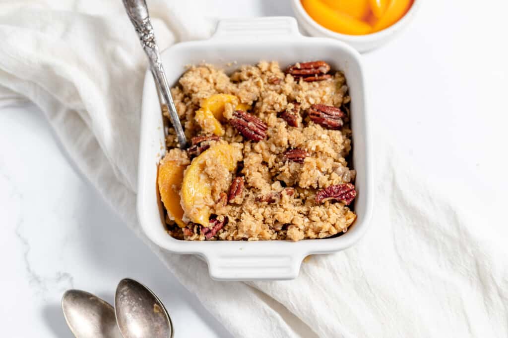 A square white dish filled with peach crisp topped with pecans sits on a white cloth, with a spoon in the dish and two additional spoons nearby.