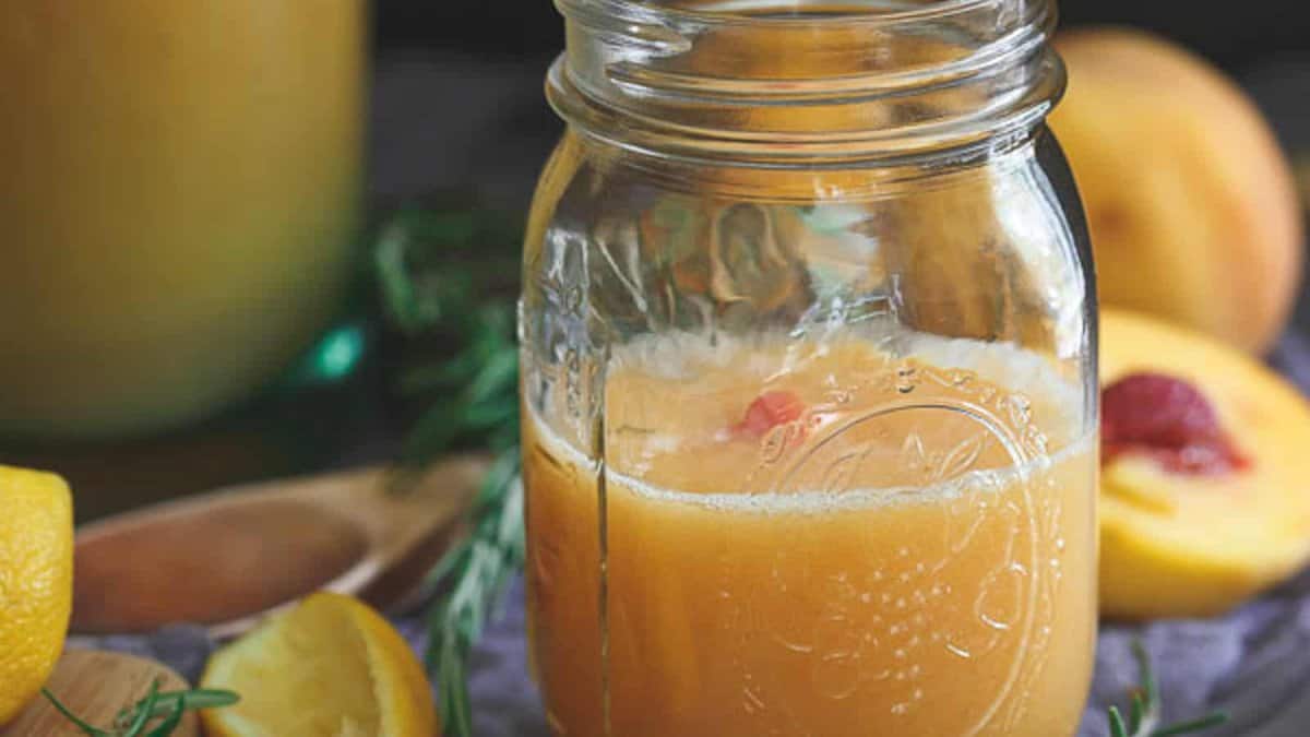 A mason jar filled with peach-colored juice sits on a table next to peach halves, lemon slices, and a wooden spoon.