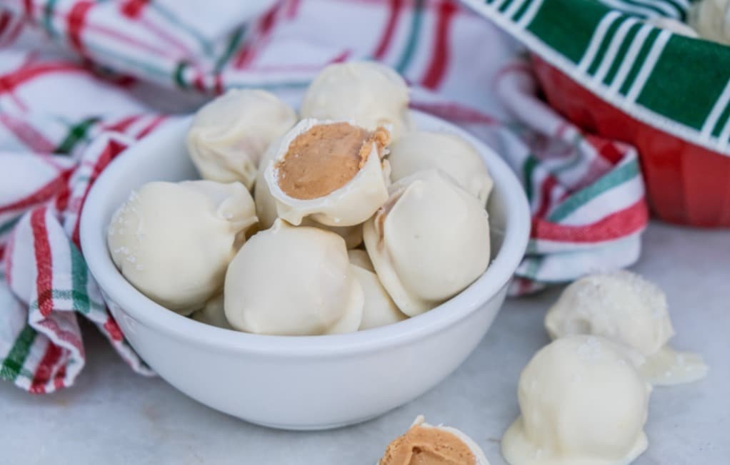 A small white bowl filled with white chocolate-coated truffles, one with a bite showing a peanut butter filling, set on a marble surface with a striped cloth in the background.
