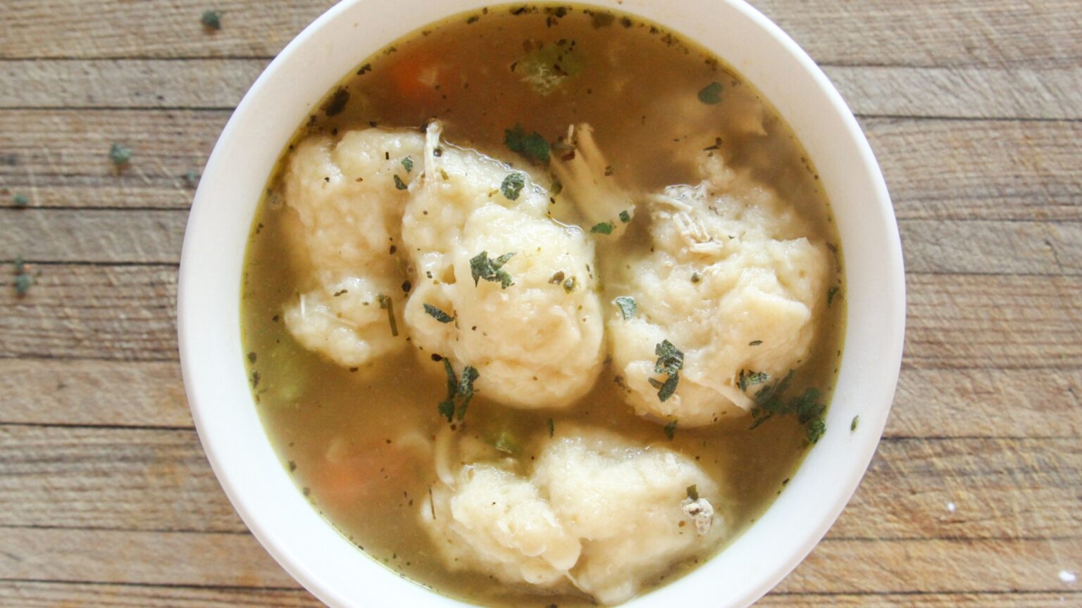 A bowl of chicken and dumplings soup with herbs, shredded chicken, and vegetables on a wooden surface.