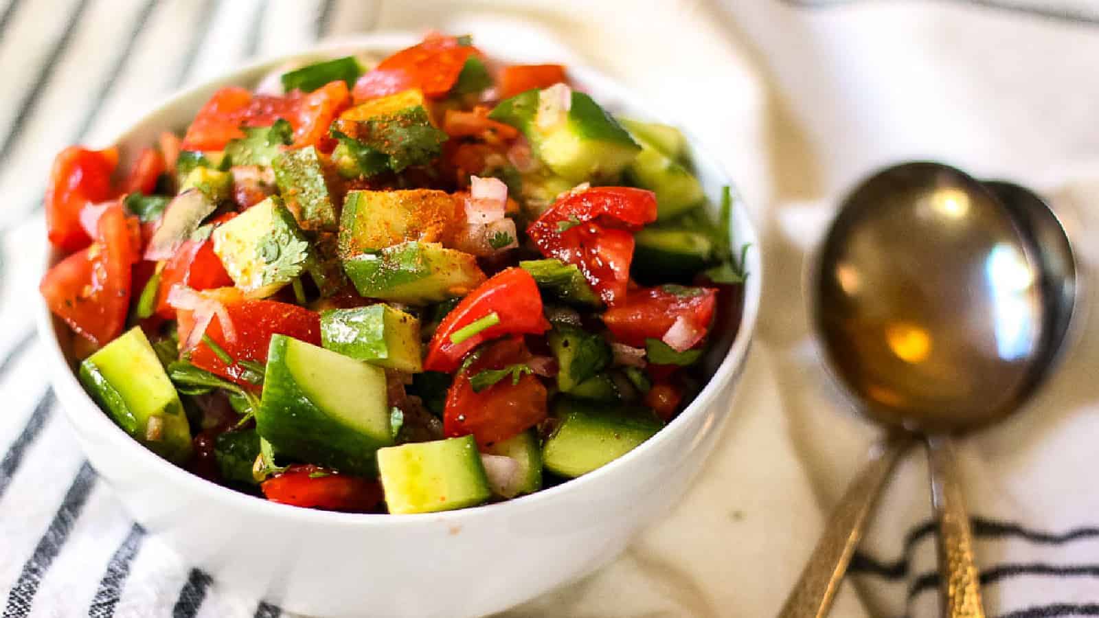 A white bowl filled with chopped tomatoes, cucumbers, avocado, onion, and cilantro sits next to two metal spoons on a striped cloth.