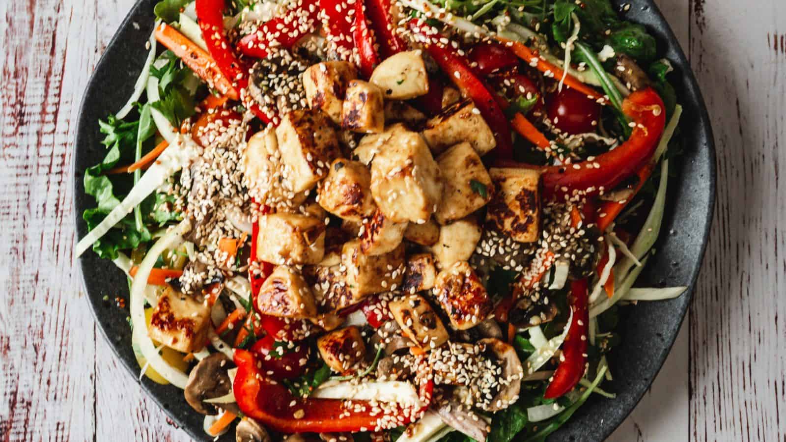 A plate of salad topped with grilled tofu, sliced red bell peppers, mushrooms, shredded vegetables, and sprinkled with sesame seeds.