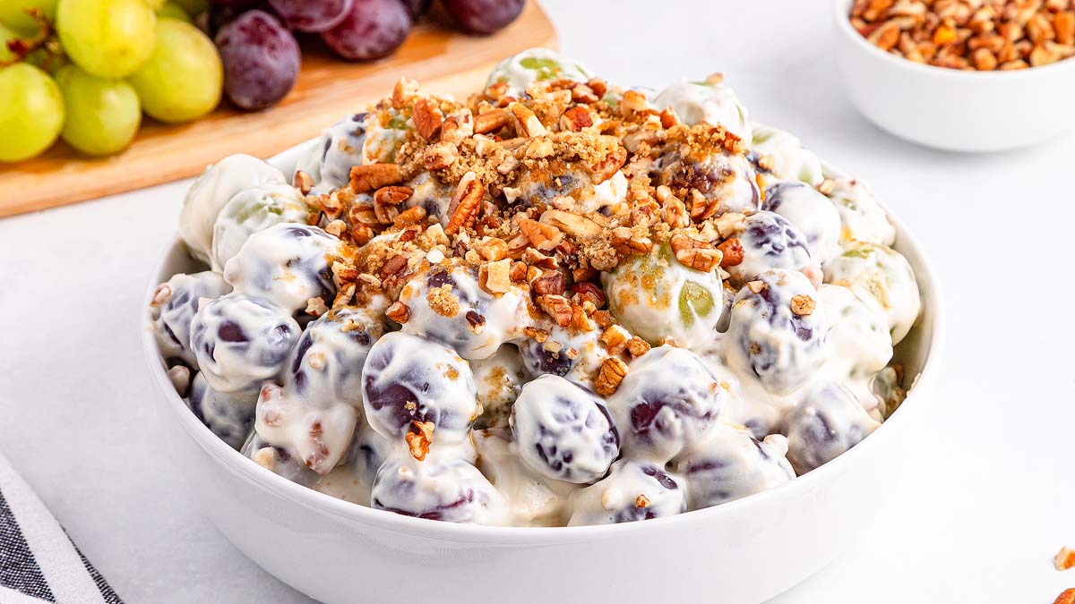 A bowl of grape salad topped with chopped nuts, with green and red grapes visible inside a creamy dressing; additional grapes and nuts are in the background.
