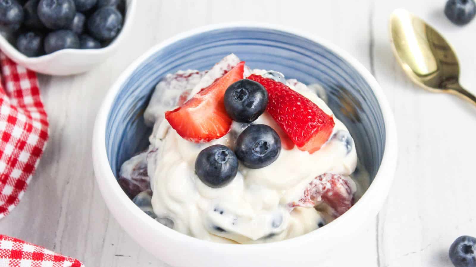 A bowl of creamy fruit salad topped with fresh blueberries and strawberry slices, placed on a white table beside a gold spoon, a small bowl of blueberries, and a red checkered napkin.