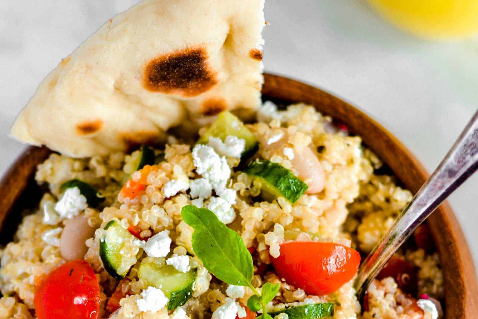 A bowl of quinoa salad with cherry tomatoes, cucumbers, white beans, crumbled feta, fresh greens, and a piece of grilled flatbread, with a spoon on the side.