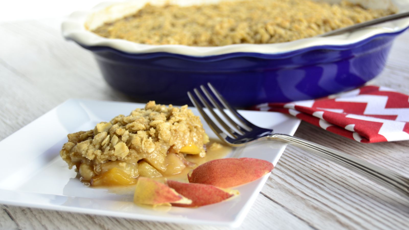A slice of peach crumble on a square white plate with a fork, peach slices, and a blue baking dish in the background.