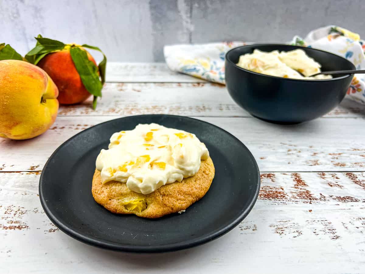 A cookie topped with white frosting on a black plate, with peaches and a bowl of more cookies and frosting in the background on a white rustic table.