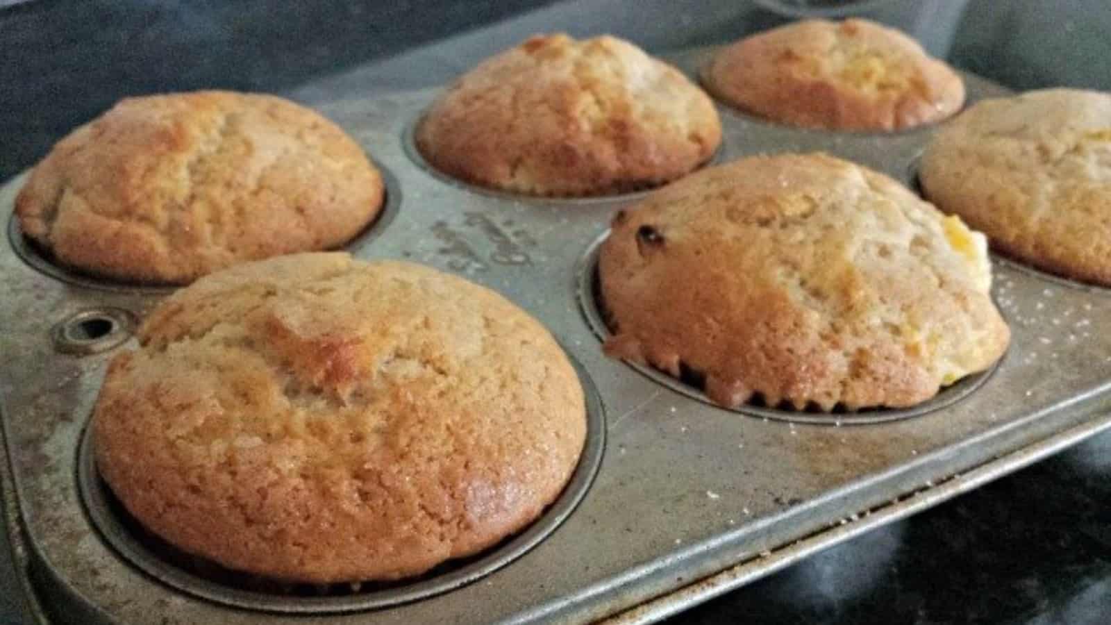 A metal muffin tin holds six freshly baked golden-brown muffins on a dark countertop.
