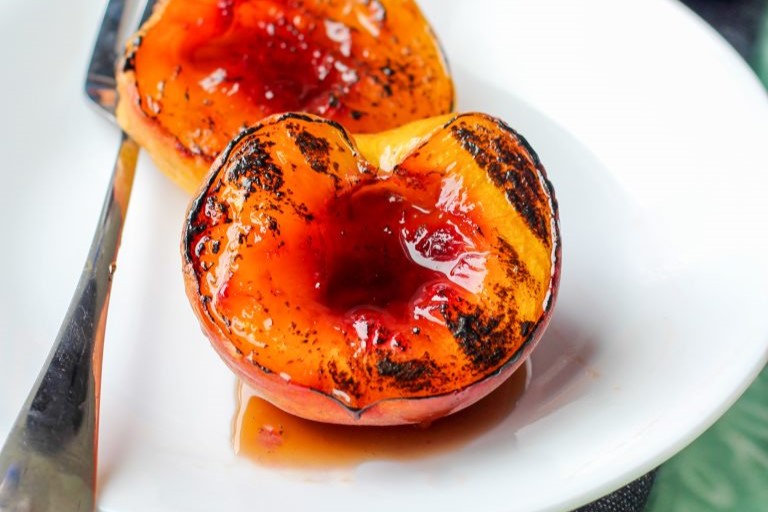 Two grilled peach halves with caramelized, slightly charred surfaces and syrupy sauce are served on a white plate with a fork.