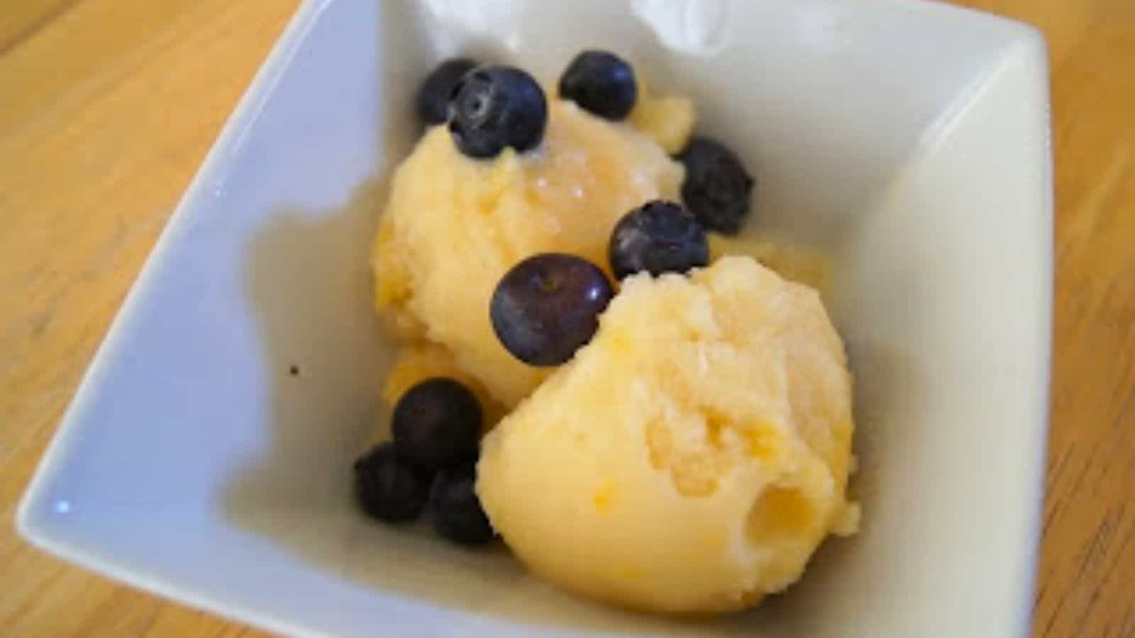 Two scoops of yellow sorbet in a white bowl, topped with several fresh blueberries, on a wooden surface.