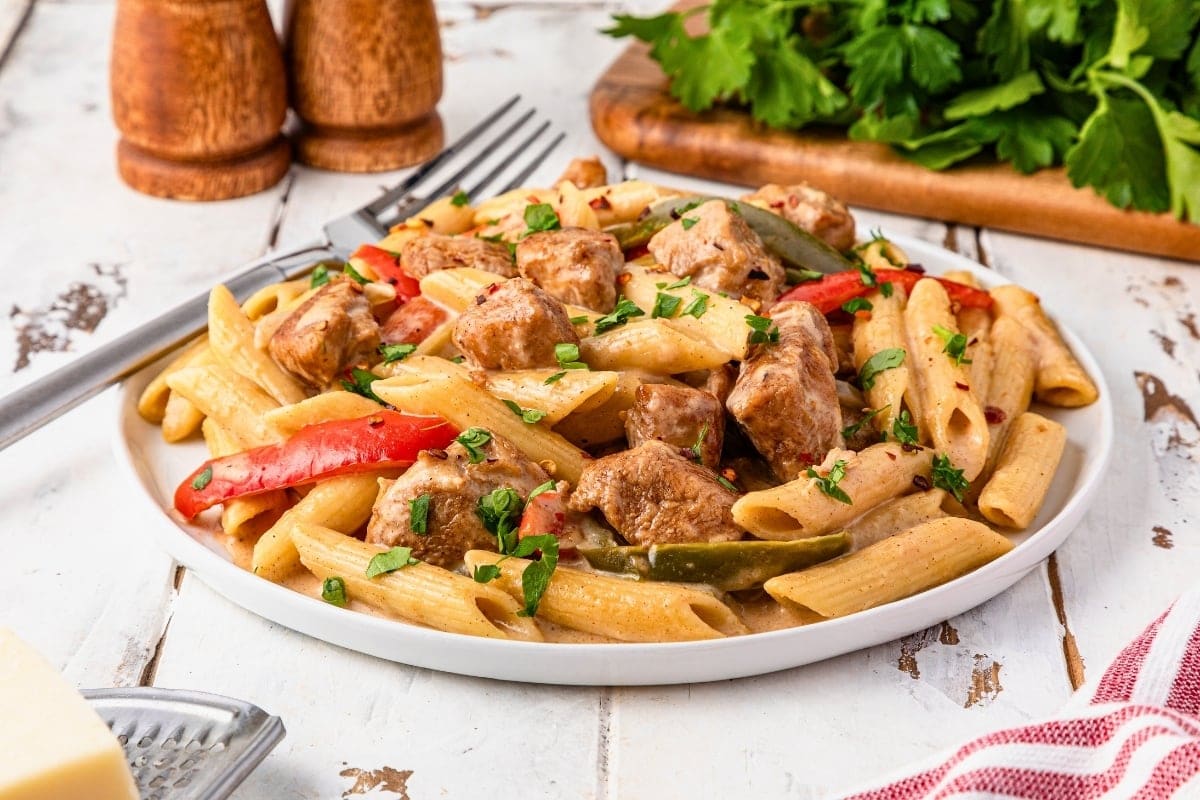 A plate of penne pasta with pieces of chicken, red and green bell peppers, and a creamy sauce, garnished with chopped herbs.