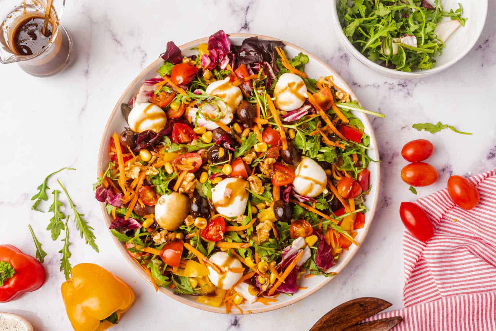 A colorful salad with mixed greens, cherry tomatoes, carrots, mushrooms, olives, and mozzarella balls, drizzled with dressing on a white plate. Nearby are salad ingredients and utensils.