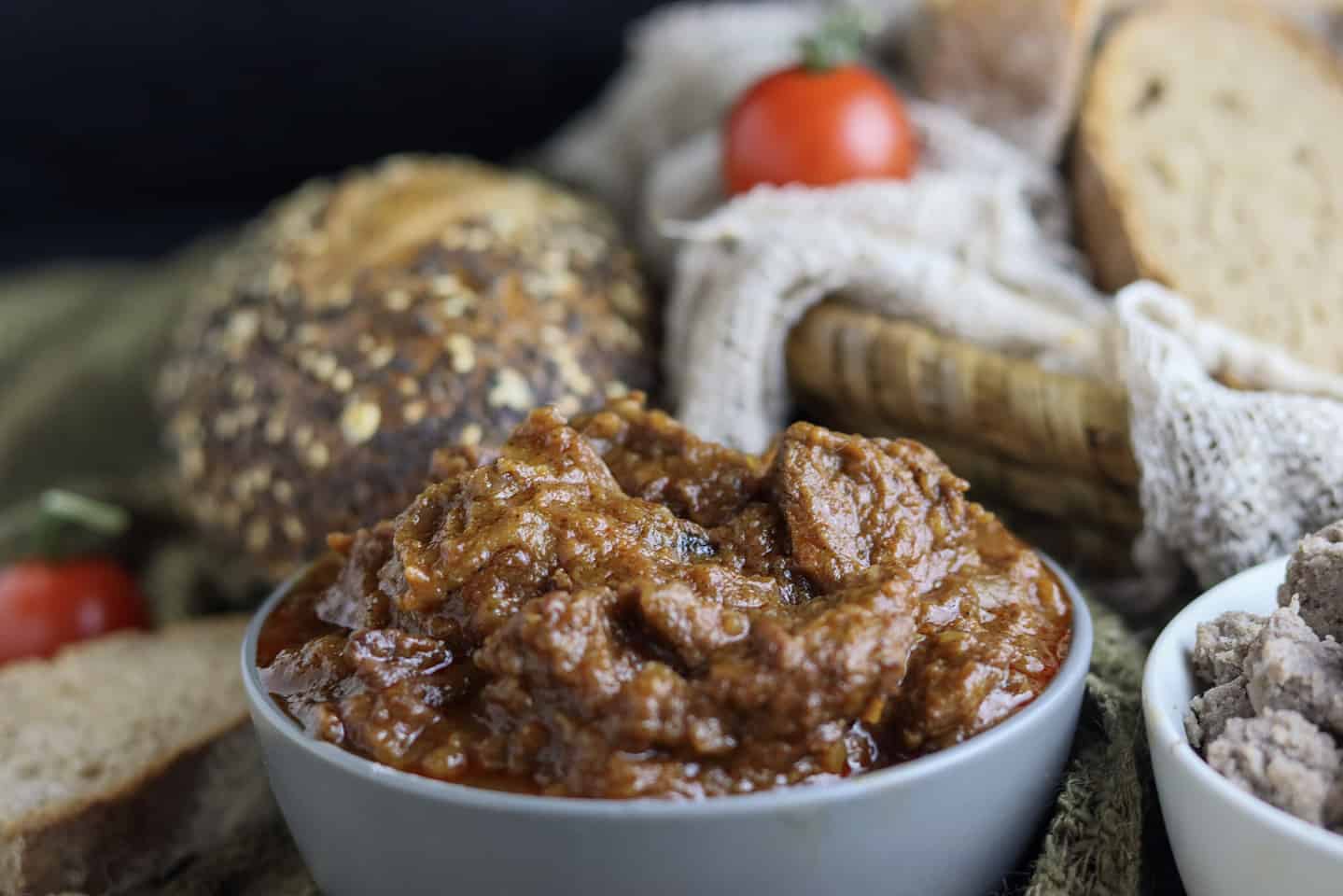 A bowl of thick, brown beef stew is in focus, with sliced bread, a whole grain roll, and a fresh tomato in the background.