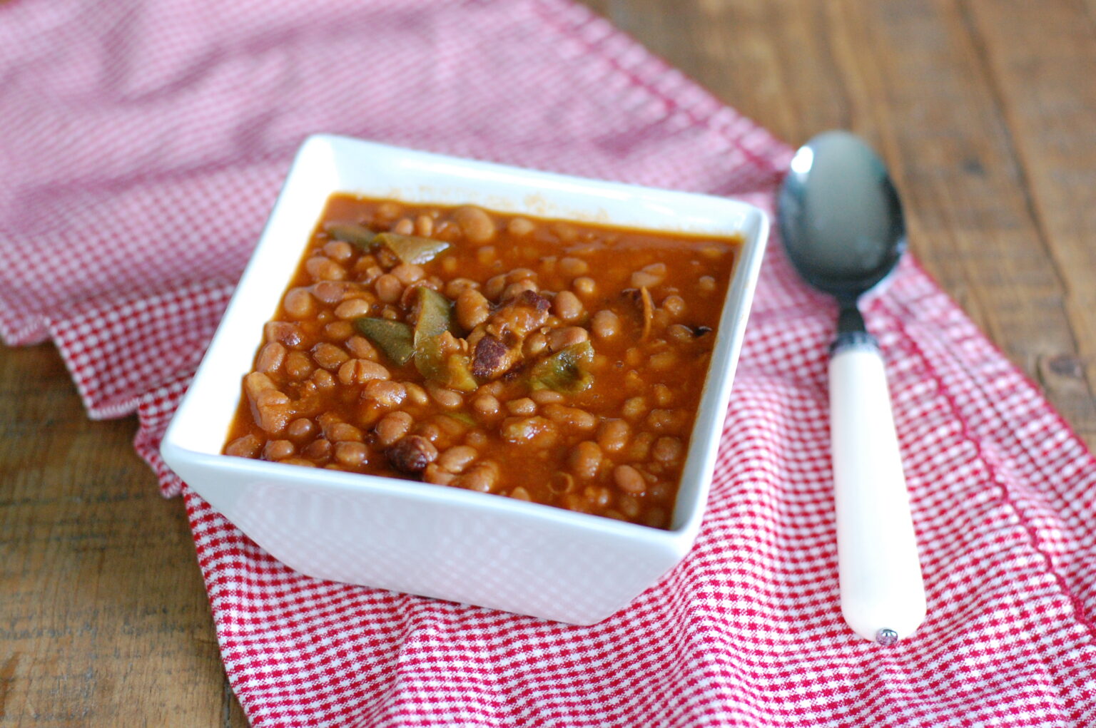 A square white bowl of bean chili sits on a red checkered cloth next to a white-handled spoon on a wooden table.
