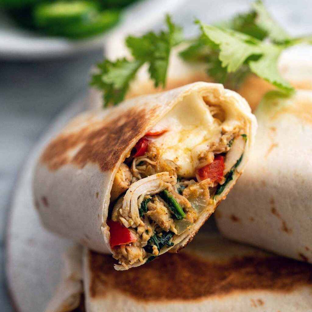 A close-up of a grilled burrito cut in half, showing chicken, red peppers, greens, and melted cheese, garnished with cilantro.