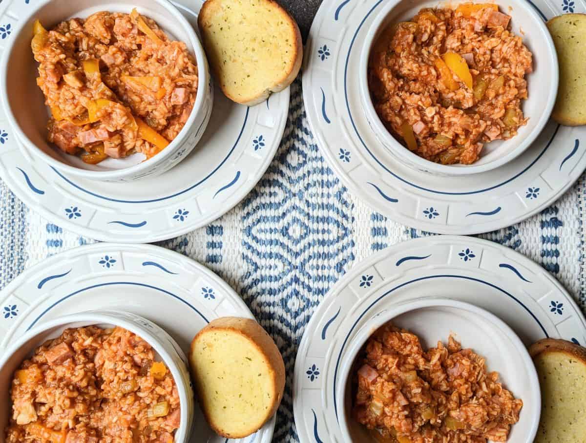 Four bowls of a tomato-based rice and vegetable dish, each served with a slice of garlic bread, arranged on patterned plates atop a woven blue and white tablecloth.