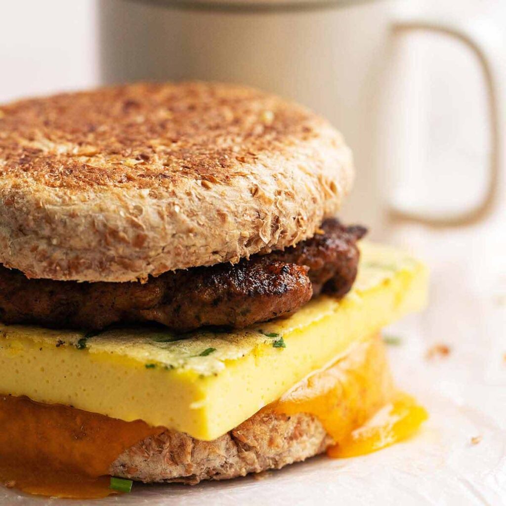 A breakfast sandwich with an English muffin, sausage patty, egg, and melted cheddar cheese, with a mug in the background.