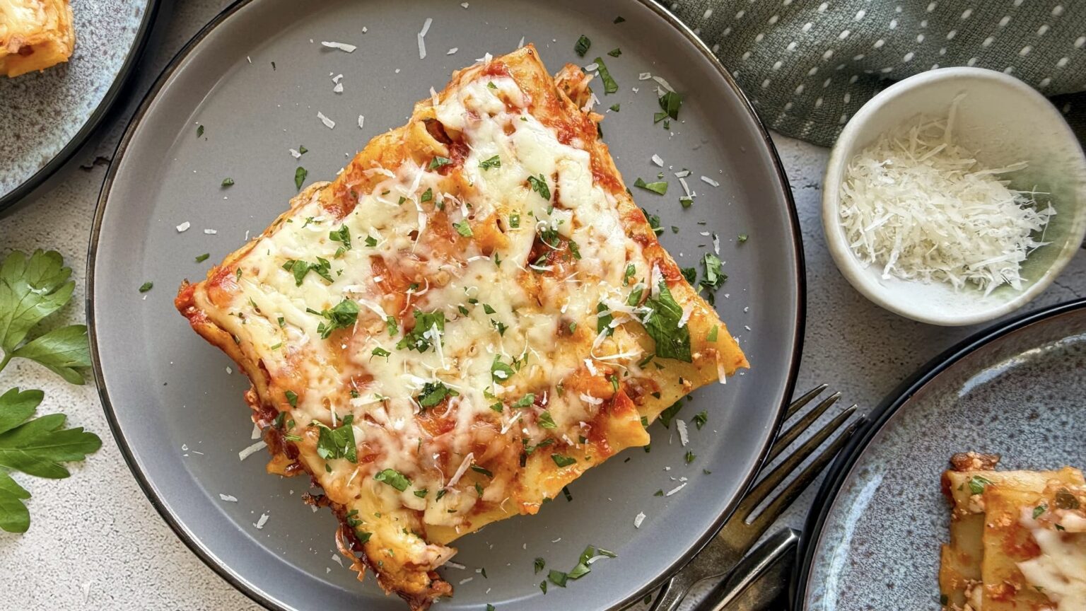 A serving of baked lasagna topped with melted cheese and chopped parsley on a gray plate, next to a small bowl of grated cheese.
