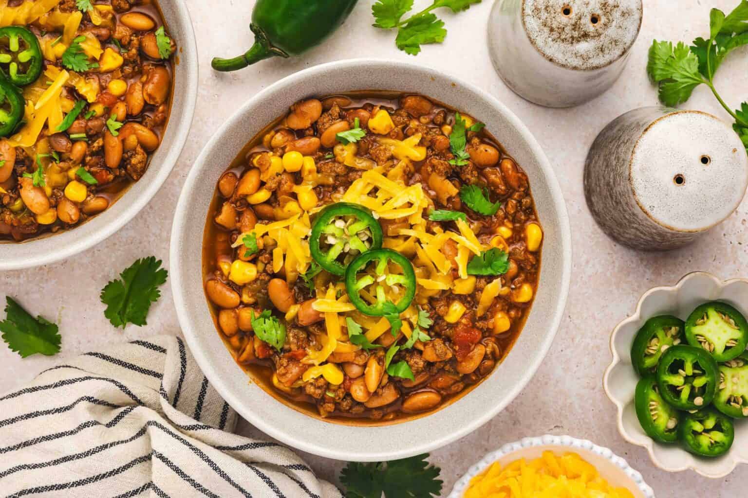 A bowl of chili with beans, corn, ground meat, shredded cheese, jalapeรฑo slices, and cilantro, surrounded by garnishes and seasonings on a light surface.