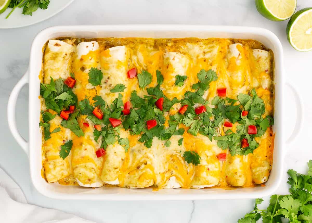 A casserole dish filled with enchiladas topped with melted cheese, chopped cilantro, and diced red peppers, on a marble surface with lime halves and herbs nearby.