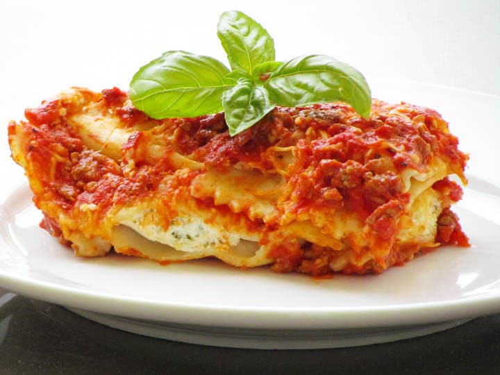A serving of lasagna with tomato sauce, meat, and ricotta cheese, garnished with a fresh basil sprig, on a white plate.