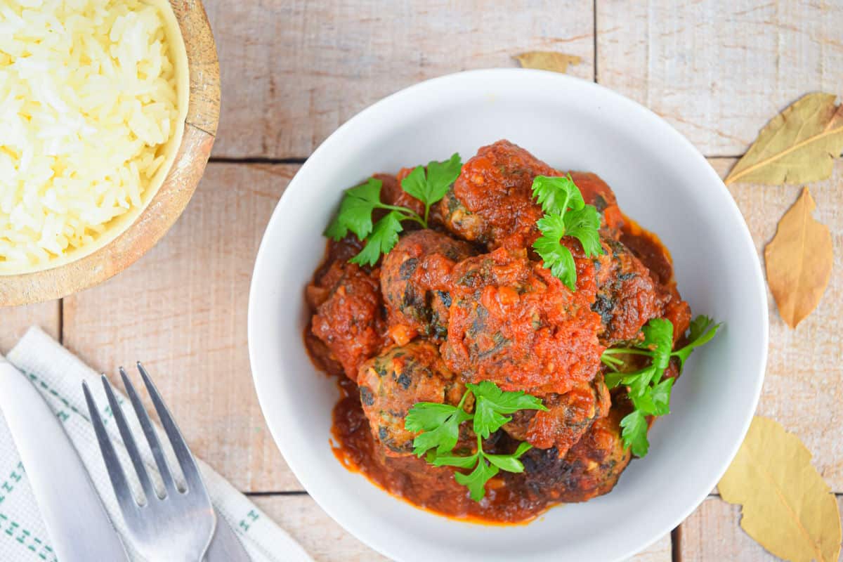 A white bowl filled with meatballs in tomato sauce, garnished with parsley, sits on a wooden surface next to a bowl of rice, a fork, and a napkin.