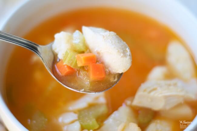 A close-up of a spoonful of chicken soup containing chicken pieces, carrots, celery, and broth, held above a bowl of the same soup.