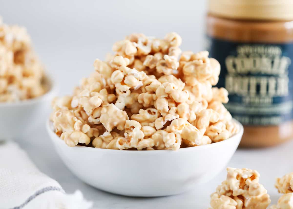 A white bowl filled with popcorn coated in a creamy topping sits on a white surface, with a jar of cookie butter blurred in the background.