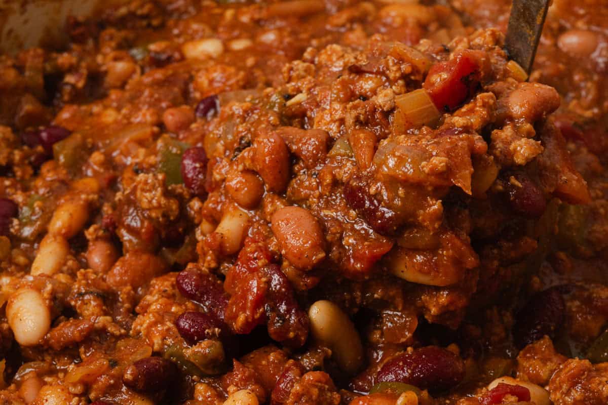 Close-up of a ladle scooping chunky chili with ground meat, beans, tomatoes, and spices.